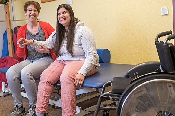 Eine Frau mit Rollstuhl und ihre Therapeutin lachen fröhlich in die Kamera