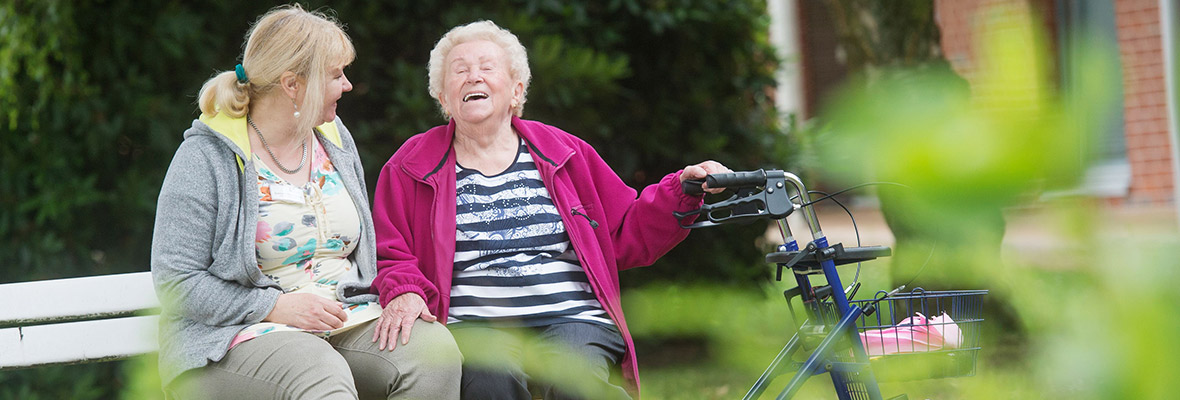 Eine alte Dame sitzt mit ihrer Pflegerin auf einer Bank und lacht